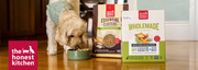 A small beige dog wearing a green top is eating from a green bowl on a wooden floor, with two bags of Honest Kitchen dog food standing beside it: Essential Clusters and Wholemade.